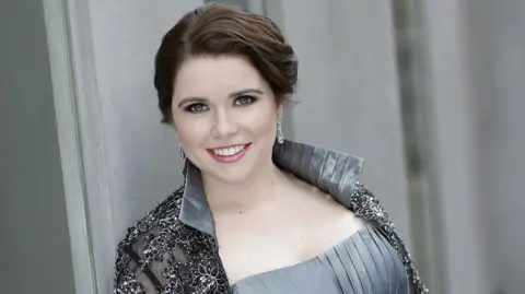 Tara Erraught stands in front of a grey wall and is smiling. She is wearing a silver formal dress and silver earrings, her hair is tied up