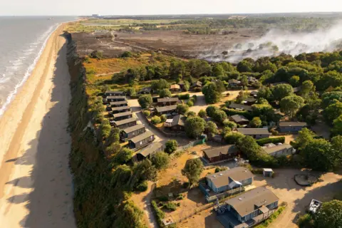 EPA/Shutterstock A bird's eye view of holiday homes, surrounded by trees, and near a cliff edge. Behind them is a smouldering black landscape. To the left is the sea.
