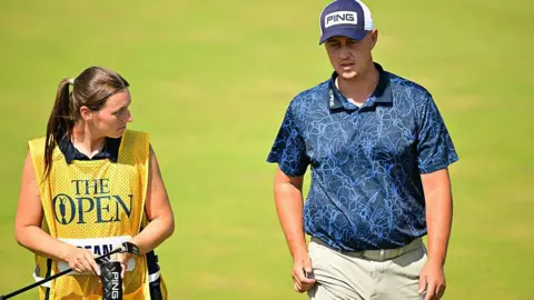 A golfer in a navy polo shirt and cap strides up the fairway of a golf course next to his female caddie.