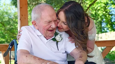 A woman in a wedding dress on the right smiles as she looks at a man. The elderly man is wearing a white roses pinned to to his top and is sitting in a wheelchair with the handle visible on the left.