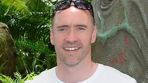 Paul Doyle has short dark greying hair with sunglasses on his head and is wearing a white T-shirt.