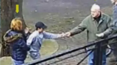 The Dawn Sturgess Inquiry CCTV showing Sergei Skripal handing bread to a young boy while they feed ducks. Sergei is in a green coat and the young boy wears a dark cap and grey top.