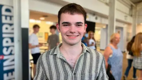Emily Doughty / BBC Stephen has short hair and wears a grey and white striped shirt. He is smiling and holding papers with his results on. In the background people are milling about in a school hall