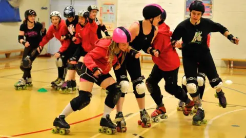 Roller derby players wearing helmets and protective gear train indoors on a sports court. Several skaters in red hoodies and black outfits form a line while others practise blocking and manoeuvres.