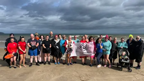 Seacoast Dippers A group of swimmers standing on a beach holding a Seacoast Dippers banner with waves behind them.