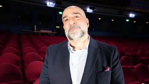 Qays Najm/BBC A wider view of Djalili standing in the Regent Theatre. He angles his head slightly away from the camera as he smiles. The red seats of the theatre are behind him.