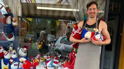 Stuart Race Mr Race is pictured holding some of the knitted hens outside his shop. He is facing the camera and smiling while wearing an overall. Other knitted hens sit in the display in his shop window.