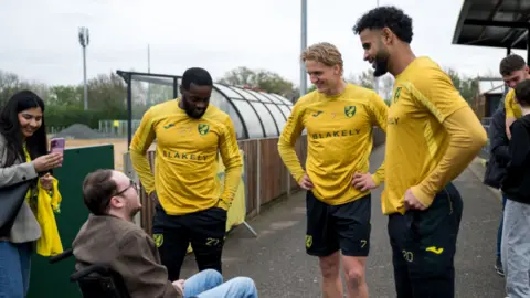 Courtney Pedlar/Norwich City FC Jonny Butcher sitting in a wheelchair chatting to three Norwich City players.