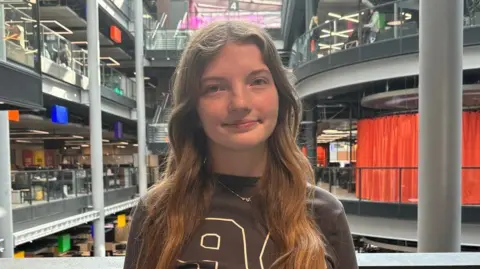 Mia is smiling at the camera. She is stood on the balcony in the BBC Wales building in Cardiff