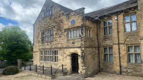 A 17th Century sandstone building with stone mullion windows and a blue plaque above a first floor window over the doorway. To the left is a mature tree and there is a cloudy sky with patches of blue.