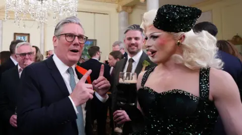 Getty Images A man in a navy suit, white shirt and green tie, talks to a man in a green sparkly dress. The man wears black gloves and sparkly green hat whilst he holds a Guinness. He has long blonde hair and makeup on. Behind them are dozens of people, out of focus in a large cream room. 