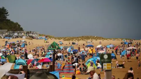 Shaun Whitmore/BBC A crowded beach with tents, windbreaks and people on it. There are grass-covered dunes and beach huts in the background.