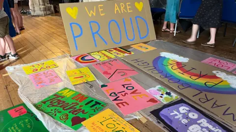 Signs with rainbows and bright colours. Examples of the words on the banners include 'love is love' and 'all in it together'.