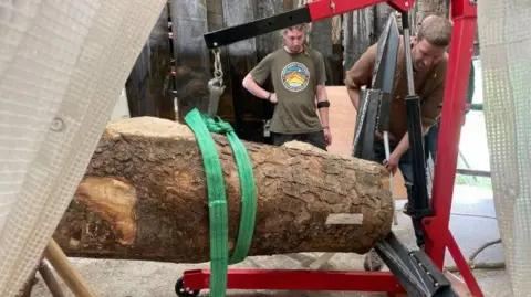 Charlie Whinney and an assistant manoeuvre the trunk with the use of a red crane. The trunk is suspended by a green strap.