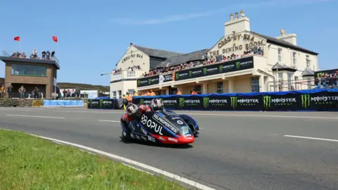 ISLE OF MAN TT A black and red sidecar outfit travelling past the Creg ny Baa, which has crash barriers in front of it.