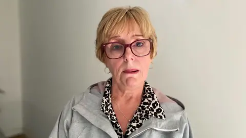 A woman looking visibly upset looks at the camera. She has dark pink-rimmed glasses and short blonde hair. She has pink lipstick on her quivering lip. She has a grey hoody on under which she has a leopard print shirt, you can see the collars of this. It is a head and shoulders shot of her.