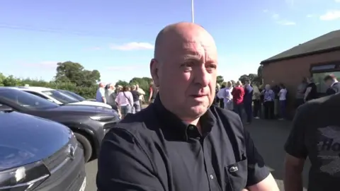 A bald man in a black shirt is standing with his arms crossed. There are black cars parked behind him on his left, a village hall on his right and people standing in front of it