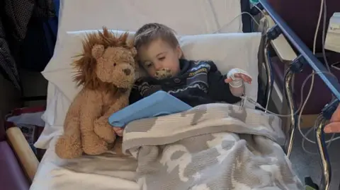 Submitted A four-year-old boy with blond hair lies in a hospital bed with his hand on a drip. He is beneath a blanket with a stuffed toy lion next to him as he looks at a tablet with a blue protective case on it.