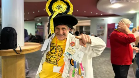 Carroll Weston/BBC Nick the flying brick wearing a tall hat with a large yellow rosette on it , a white coat and a yellow T-shirt points at the camera 