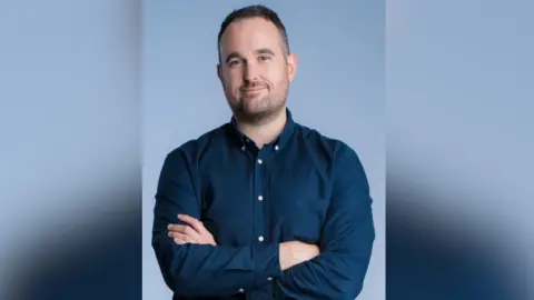 Presseye Richard Morgan, a man with short dark hair in a navy shirt, with his arms folded