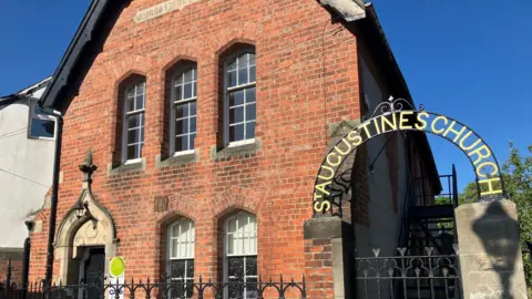 A red brick old style building, formerly a school now used as a church hall. it is made of red brick with five large windows. An arch can be seen with the lettering St Augustines Church