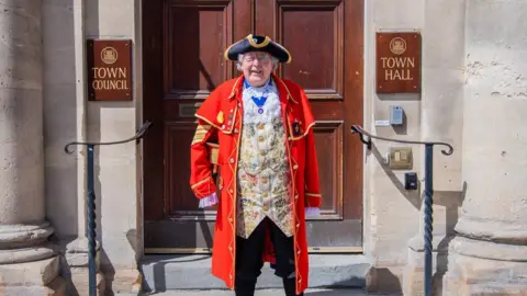 Jack Boskett An older gentleman town crier stands in front of the entrance to the town hall wearing traditional town crier clothes. He is smiling in the sunshine.