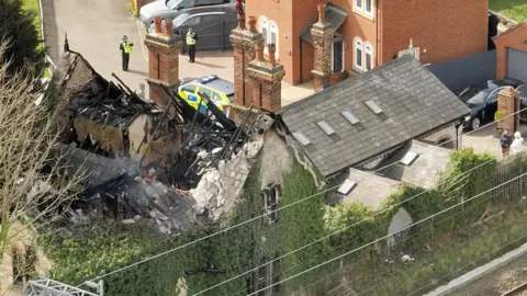 An overhead look at a building with a burned out roof, with blackened timbers, next to a railway line. Two police officers are standing in the driveway, with a police car just seen through the building's chimneys. 