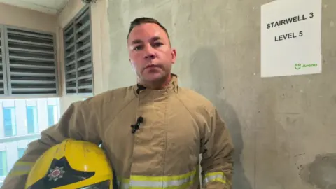 Fireman Jamie Oldfield holds his yellow fire helmet while wearing his brown uniform