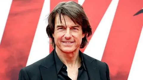 Tom Cruise attends the Mission: Impossible – The Final Reckoning premiere in New York, earlier this year. He is standing in front of a promotional sign for the film, wearing a black shirt and black jacket and smiling at the camera.