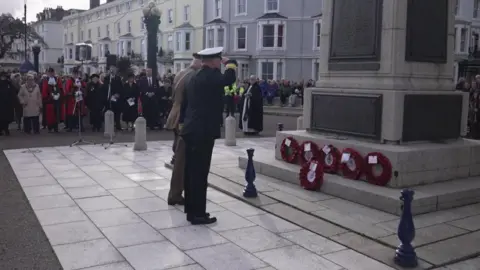 The man is seen standing before wreaths of poppy's with his arm raised to salute. He is stood next to another man taking part in the ceremony as on lookings in the town gather to watch.