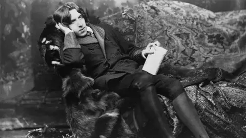 Oscar Wilde lounging on a chair with paper in his hand in a black and white portrait.