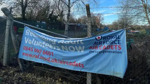 An air cadet recruitment banner on a fence outside the site, which has a small wooden building in the distance.