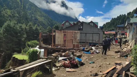 Khumbu Pashanlhamu 농촌 자치구 사람들이 부분적으로 무너진 언덕 옆의 잔해와 파괴된 건물들 사이로 비포장 도로를 걷고 있습니다.