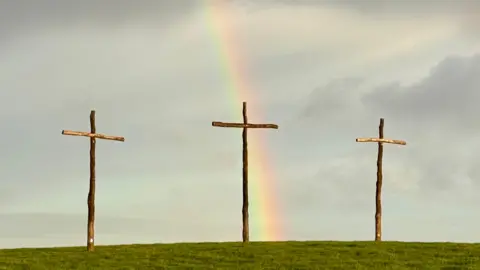 rainbow catcher Three wooden crosses on a grassy hill, with the curved arch of a rainbow behind.