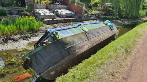 Robin Lowbridge A long boat with a pale green upper area lying on its side in a canal with very little water in it and gardens leading down to the canal on the far side
