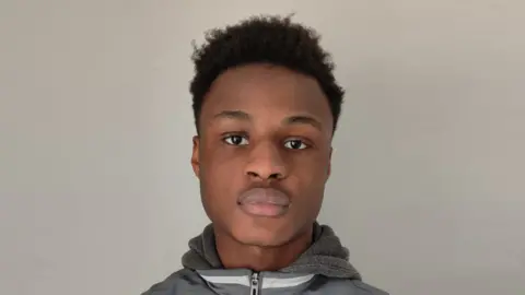A man with dark hair, wearing a hoodie, pictured close-up to his face with a plain white wall behind him