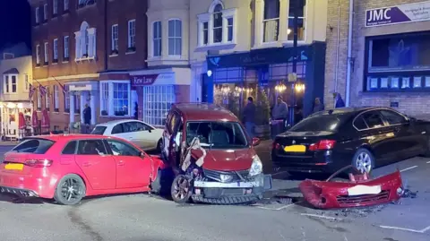 A collision between two vehicles on a high street. A light red coloured Audi is shown in the side of a darker red coloured vehicle which appears to have suffered the more significant damage. It's bumper is fully detached and airbags seem to have deployed in both cars. A group of bystanders are standing nearby and surveying the scene