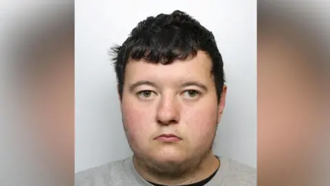 A man photographed against a plain, light grey background, stares at the camera. He has dark, short, and slightly curly hair and is wearing a light grey crew‑neck sweatshirt.