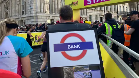 Nicky Ford/ BBC A participant runs the race while wearing a large costume of a London Underground "Bank" station sign on their back.