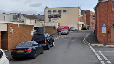 Google Google street image of Church Street in Exmouth. It shows cars parked on the left hand side of the street. Buildings line each side of the road. The sky is grey.