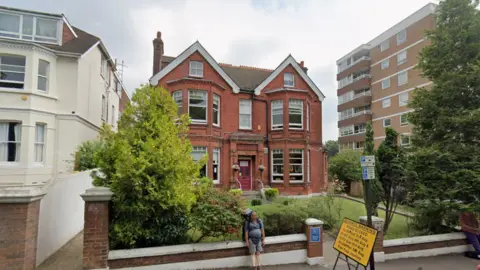 Google A red brick building. It has trees in the garden. 