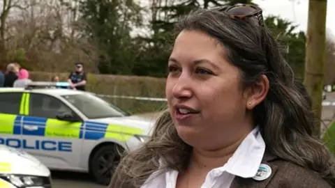Genevieve has long brown hair and sunglasses on her head. She is wearing a brown jacket and a white shirt. Police cars block the road behind her. The road is also cordoned off by an officer who stands by blue and white tape.