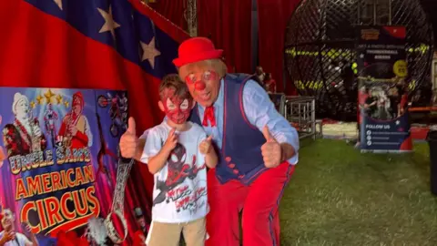 Charles A young boy with Spiderman face paint poses next to a clown with a red nose and a red hat. They stand together with the thumbs up at the camera. 