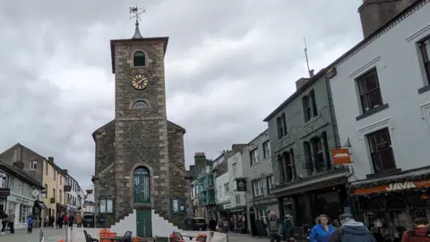 A general view of Moot Hall in Keswick. It is in the middle of a pedestrian area and has a turret with two sets of steep steps in front of it, each leading up to the central doorway. The brick building also has a black clock with golden numbers and features. There are black and orange table and chairs from a cafe outside the building.