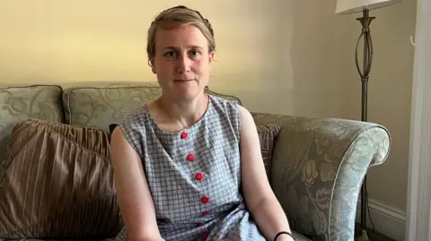 A woman sits on a green patterned sofa with brown cushions to her left. She is wearing a blue dress with red buttons. Her blonde hair is braided and tied behind her neck. 