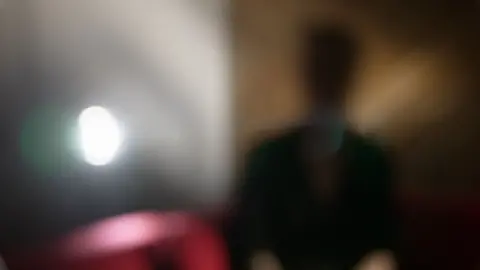 Emma sits indoors, deliberately photographed heavily out of focus so that her face and identifying features cannot be seen. A bright light is visible to the left and a red sofa is behind her.