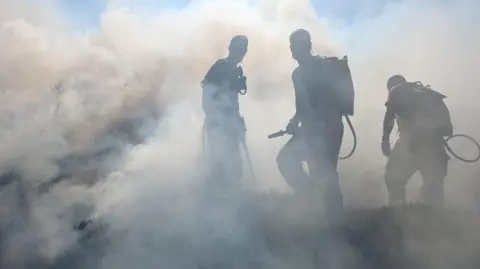 Firefighters wearing backpack water sprayers work through thick smoke while tackling a vegetation fire on open moorland. The figures are silhouetted against dense grey and white smoke that fills most of the scene.