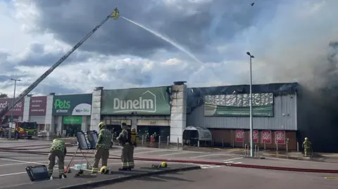 Retail park showing various units including Dunelm and Pets at Homes. The charred interior of the Dunelm shop is visible. One fire crew is directing a jet of water from above down into the smoke-filled Dunelm unit. Some firefighters are looking at a plan of the site in the foreground