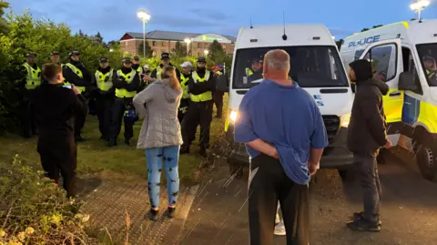 Three men and a woman have their backs to the camera as they face several police officers and vehicles outside the hotel