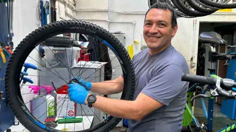 A dark-haired man in a blue T-shirt, smiling and holding a bike wheel in a workshop.